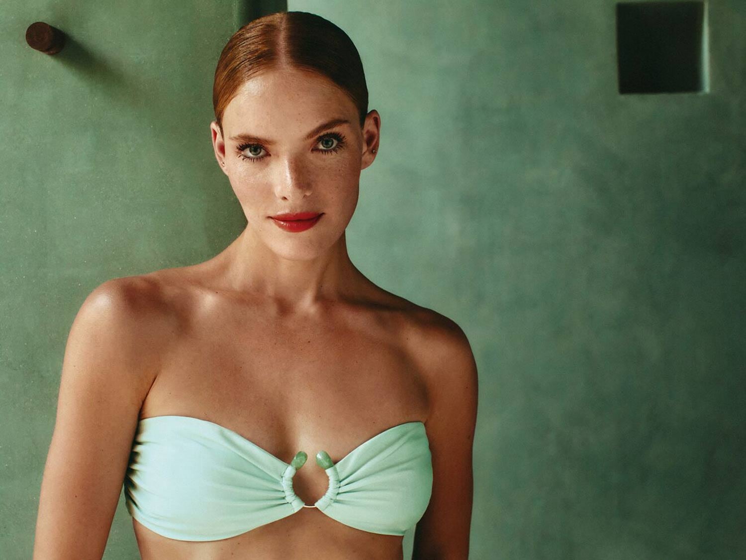 Portrait d'une femme rousse portant un haut de bikini turquoise avec des lèvres rouge vif, sur un fond vert d'eau.