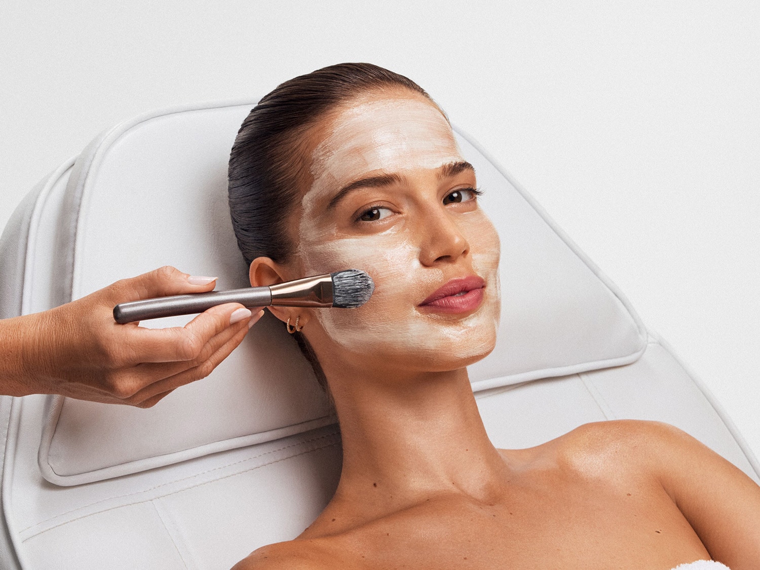 Une femme reçoit un masque facial blanc crémeux appliqué avec un pinceau gris à poils noirs, assise sur une chaise blanche.