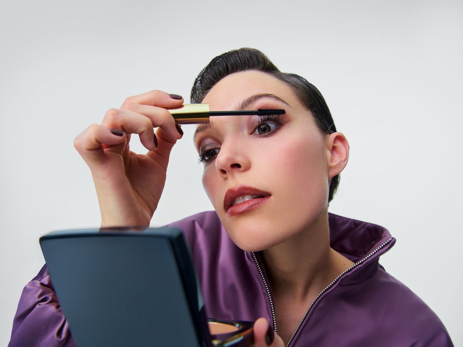 Een vrouw met donker haar en een paarse jas brengt mascara aan op haar wimpers, terwijl ze in een compact spiegeltje kijkt.