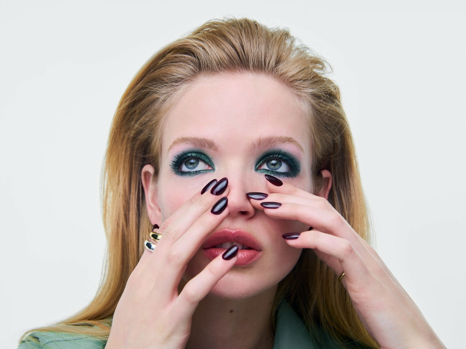 Een vrouw met lang blond haar en een groene blouse draagt donkergroene oogschaduw, donkerrode lippenstift en donkerrode nagellak, terwijl ze haar gezicht aanraakt met beide handen.