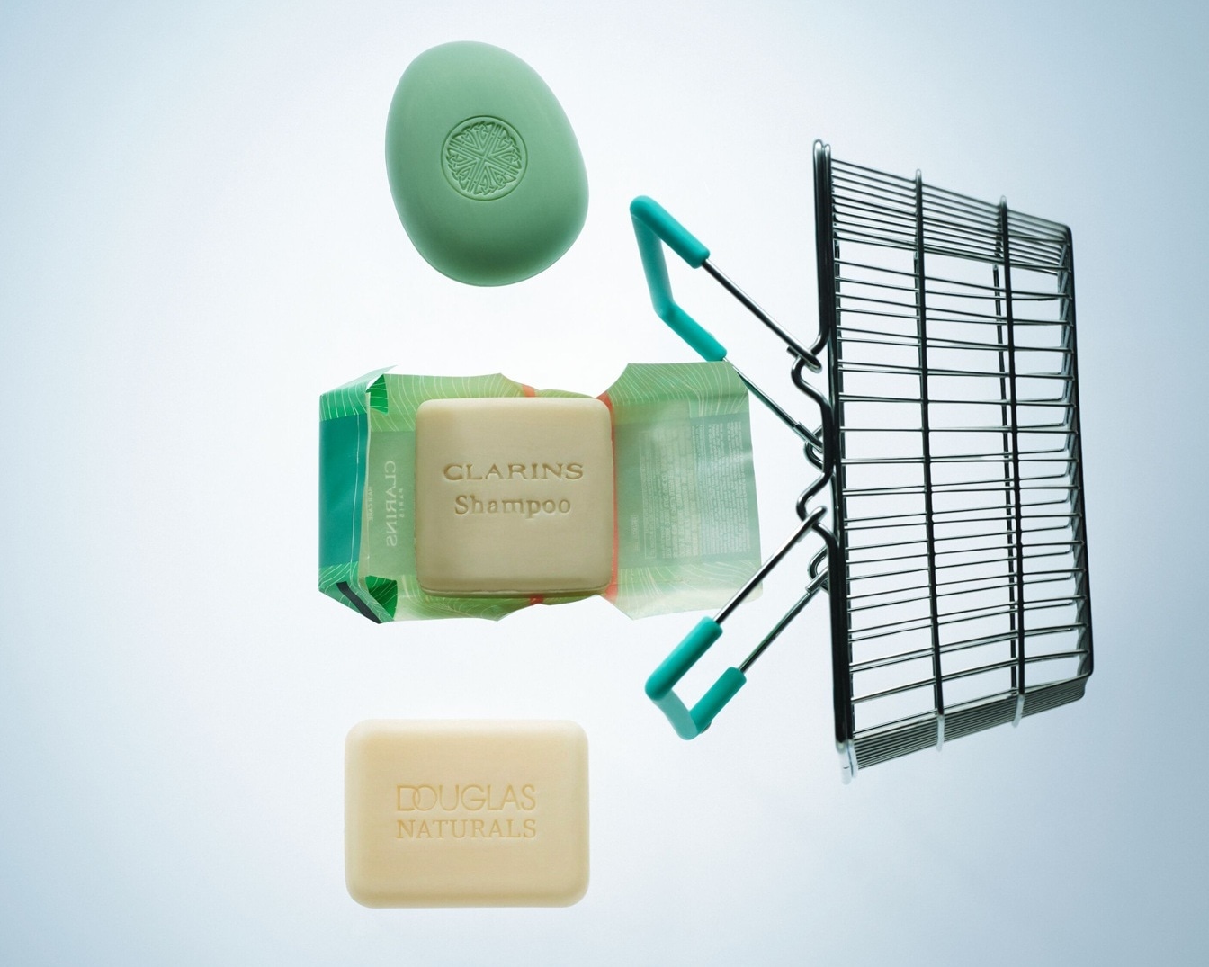 Verschiedene Seifenstücke von Marken wie Clarins und Douglas Naturals schweben neben einem Miniatur-Einkaufswagen.