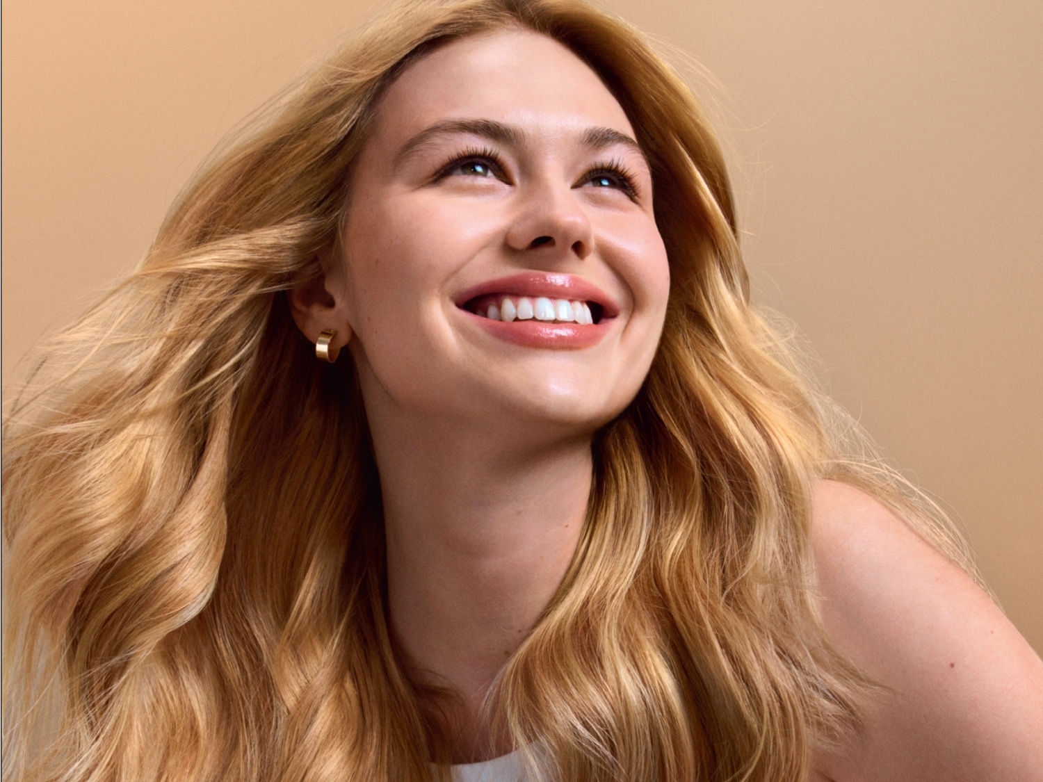 Une jeune femme blonde rayonnante, les cheveux ondulés et brillants, sourit en regardant vers le haut, avec un fond beige uni.