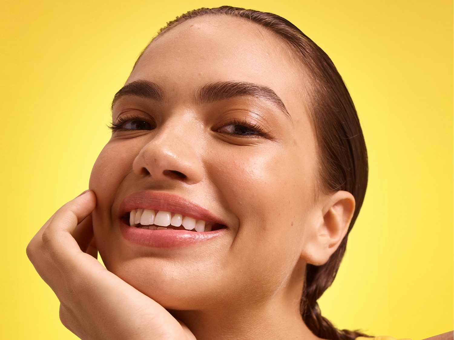 Sur un fond jaune vif, une femme rayonnante sourit, sa main délicatement posée sur sa joue, mettant en valeur son teint éclatant et ses lèvres brillantes.