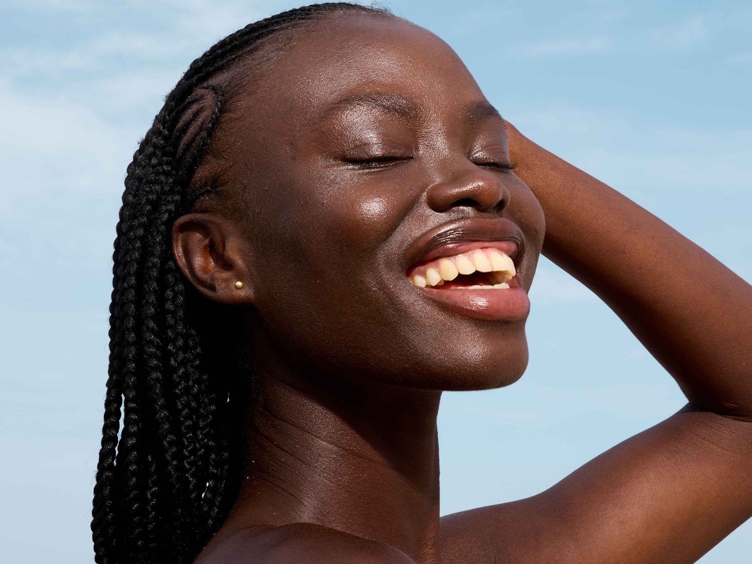 Une femme noire souriante, les yeux fermés, les cheveux tressés, la peau brillante sous le soleil, exprimant la joie et la beauté naturelle.