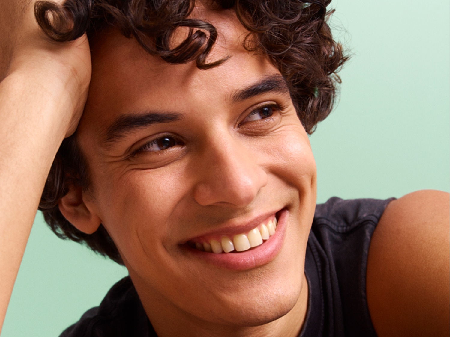Gros plan sur un jeune homme souriant aux cheveux bouclés bruns, la main sur la tête, sur un fond vert clair.