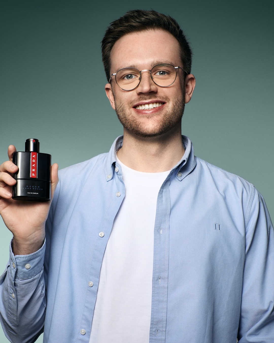 Jaunas vyras su akiniais ir šviesiai mėlynais marškiniais šypsosi ir laiko juodą Prada Luna Rossa Ocean Eau de Parfum buteliuką.