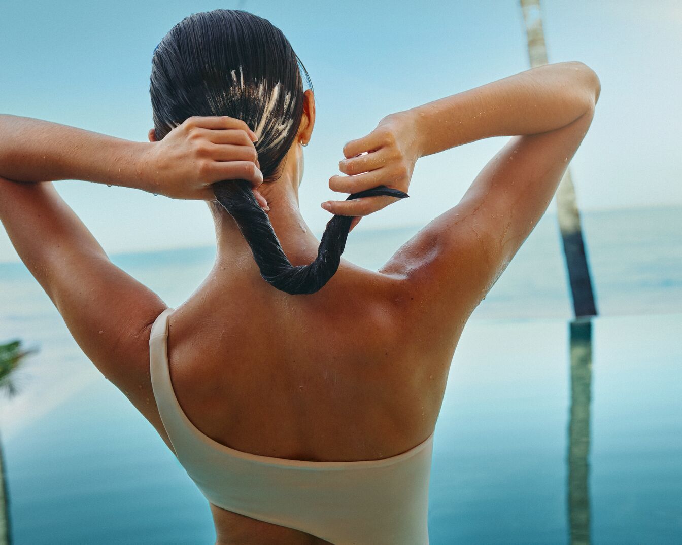 Eine Frau mit nassen, dunklen Haaren steht vor einem Pool und hält ihre Haare zu einem Zopf zusammengebunden.