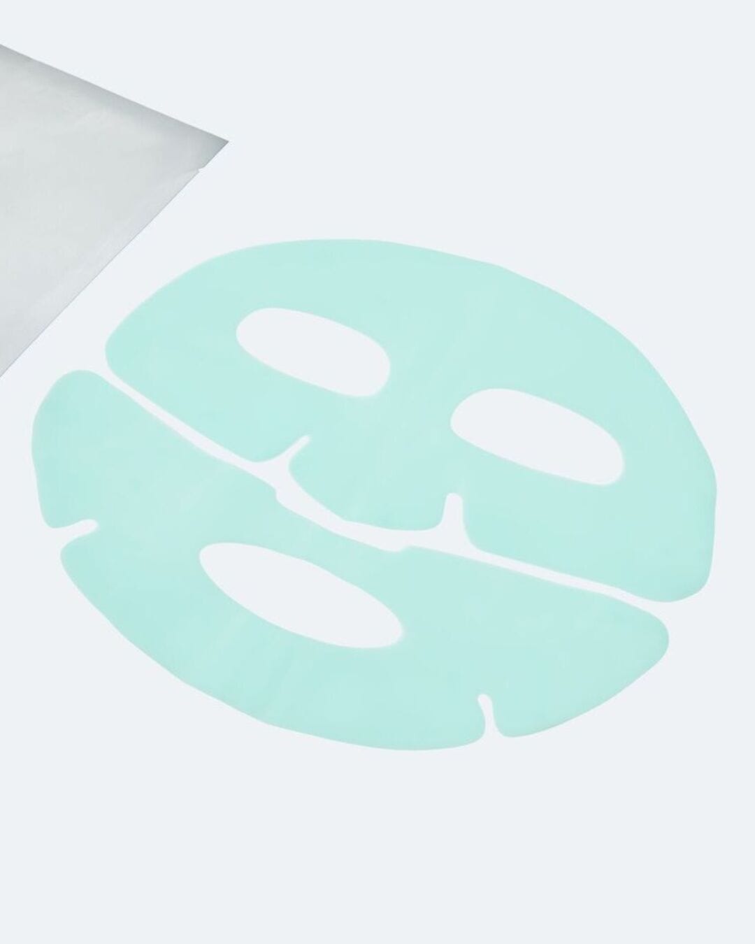 Voici un masque facial en tissu bleu clair, découpé pour les yeux, le nez et la bouche, posé sur une surface blanche à côté de son emballage argenté.