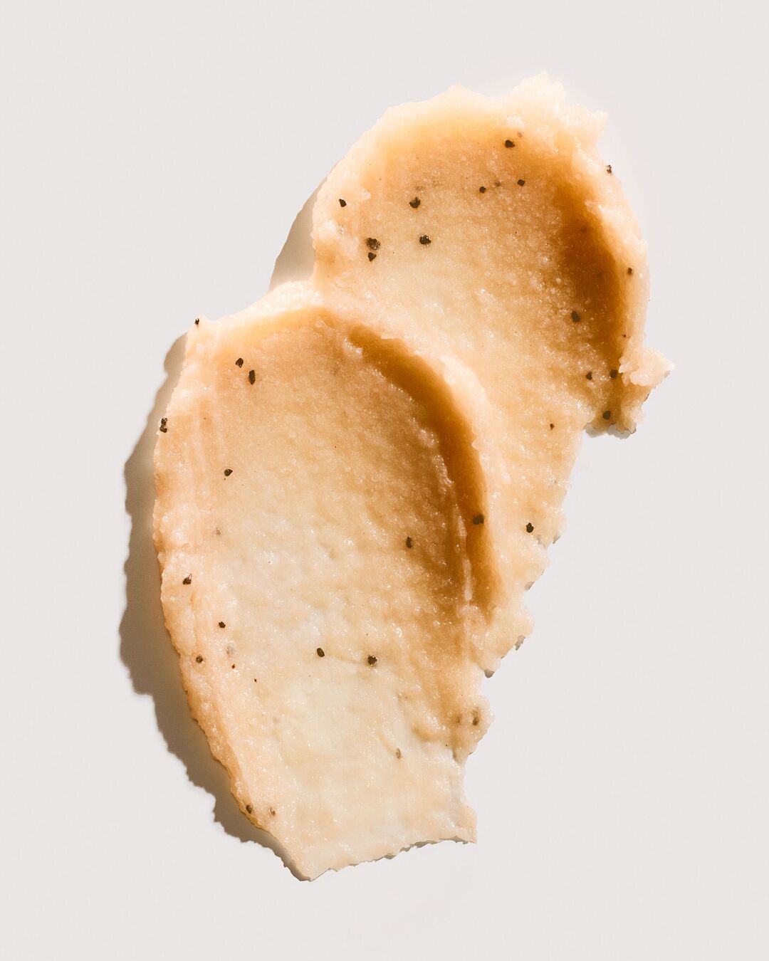 Gros plan sur un échantillon de gommage beige clair avec de fines particules exfoliantes noires, offrant une texture riche et crémeuse pour une peau douce et renouvelée.