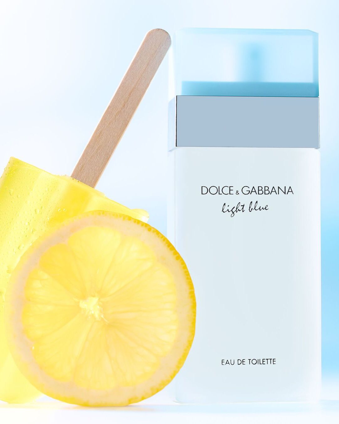 Šalia „Dolce & Gabbana Light Blue“ tualetinio vandens buteliuko pavaizduotas citrinos griežinėlis ir geltonas ledų gabalėlis.