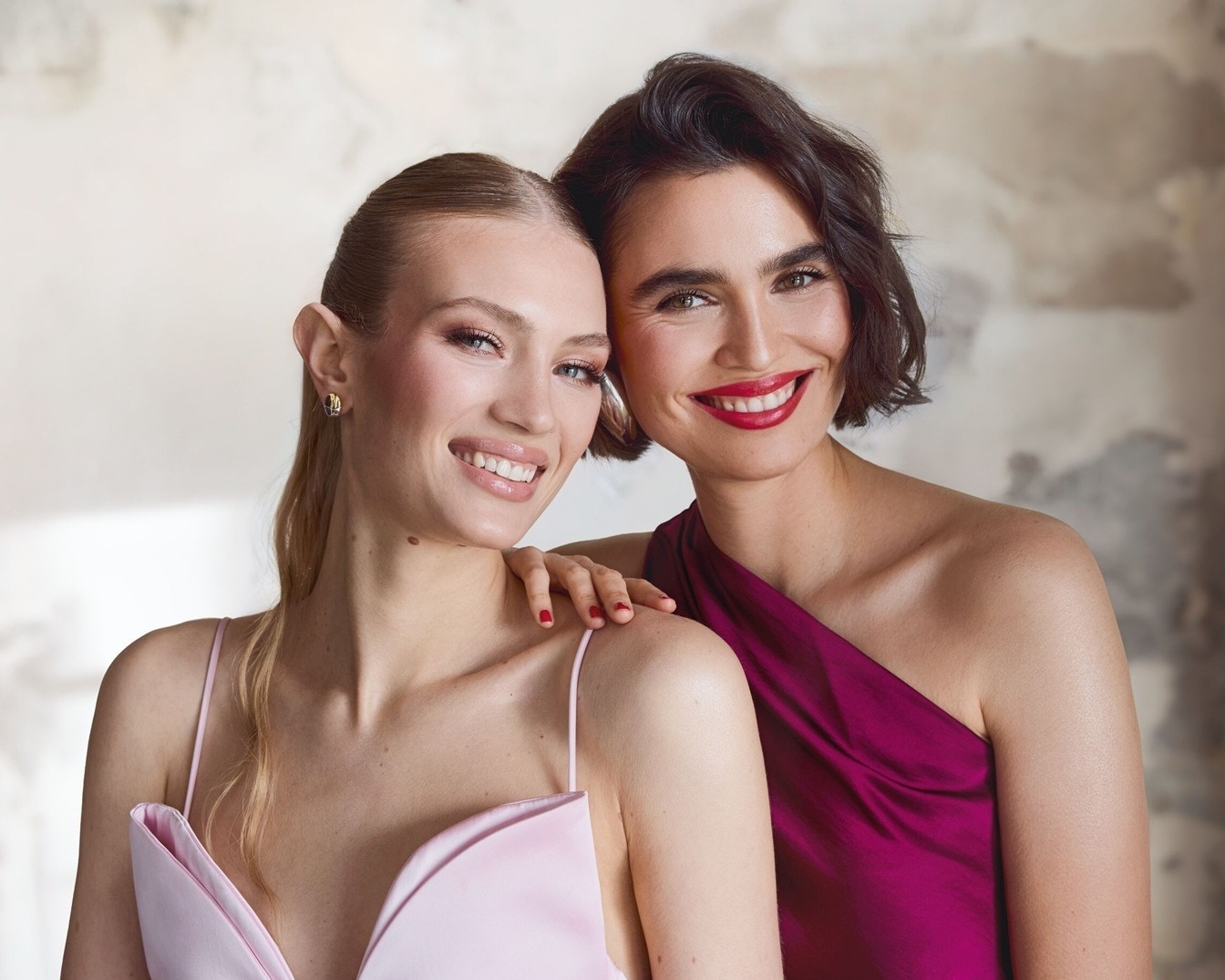 Twee lachende vrouwen, één in een roze jurk en de ander in een bordeauxrode jurk, poseren samen met rode lippenstift en een stralende glimlach.