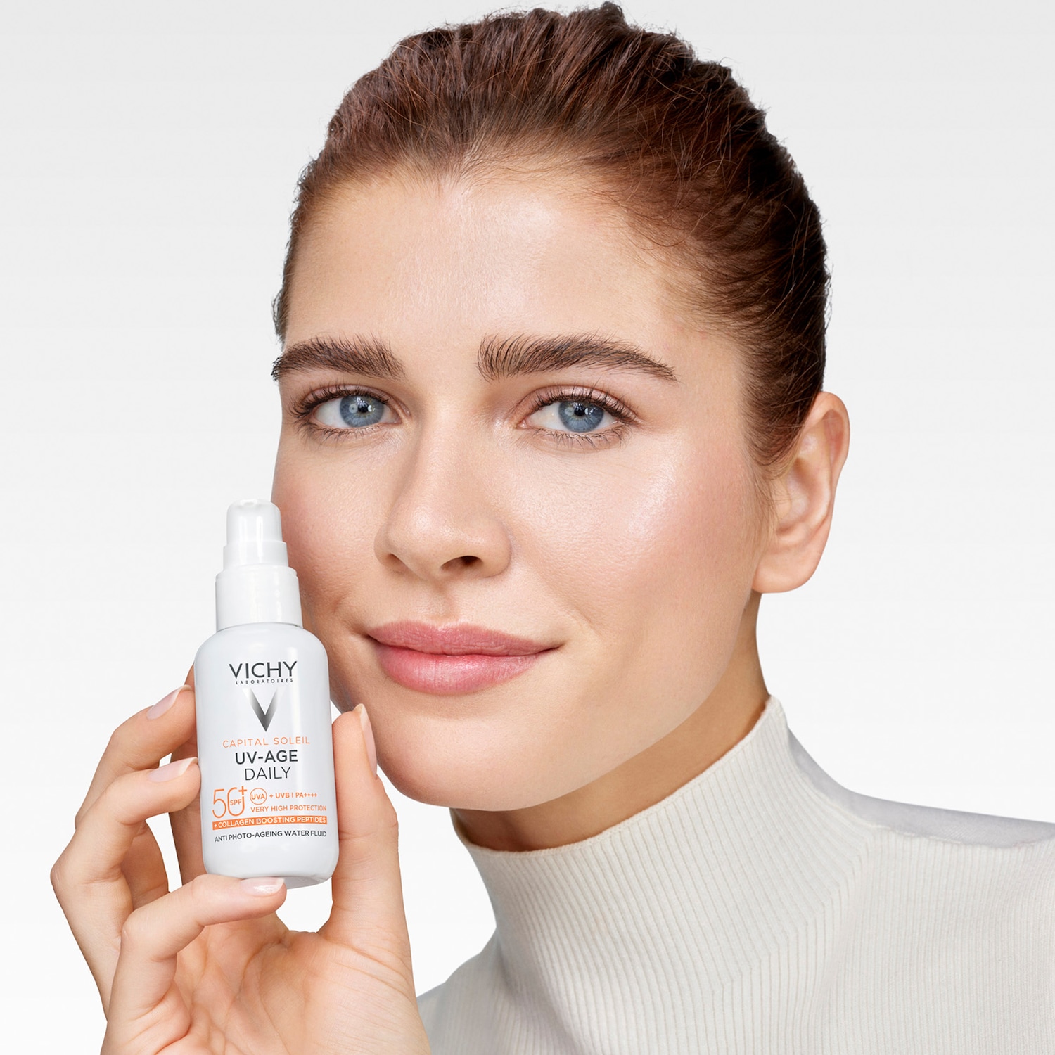 Une femme souriante tient un flacon blanc de Capital Soleil UV-Age Daily de Vichy, un fluide aqueux anti-âge avec FPS 50, offrant une très haute protection et contenant des peptides stimulant le collagène.