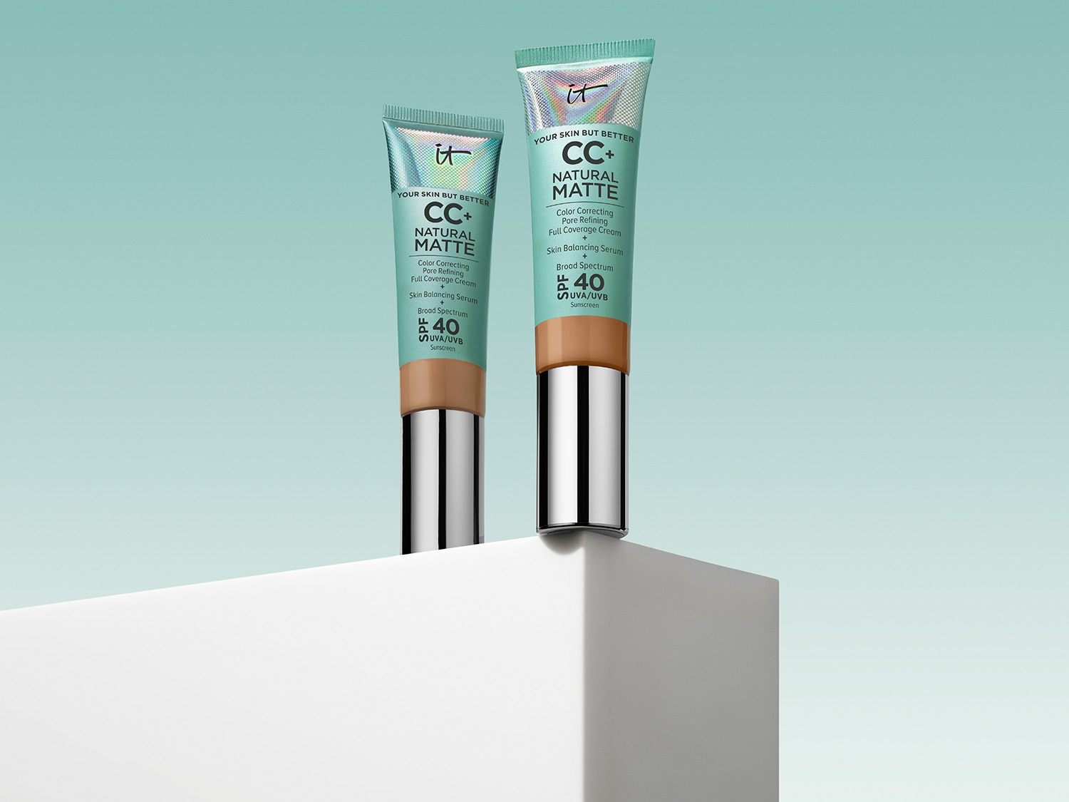Dwa opakowania kremu IT Cosmetics CC+ Natural Matte z SPF 40, obiecującego korekcję koloru, zwężenie porów i pełne krycie, stojące na białym podeście.