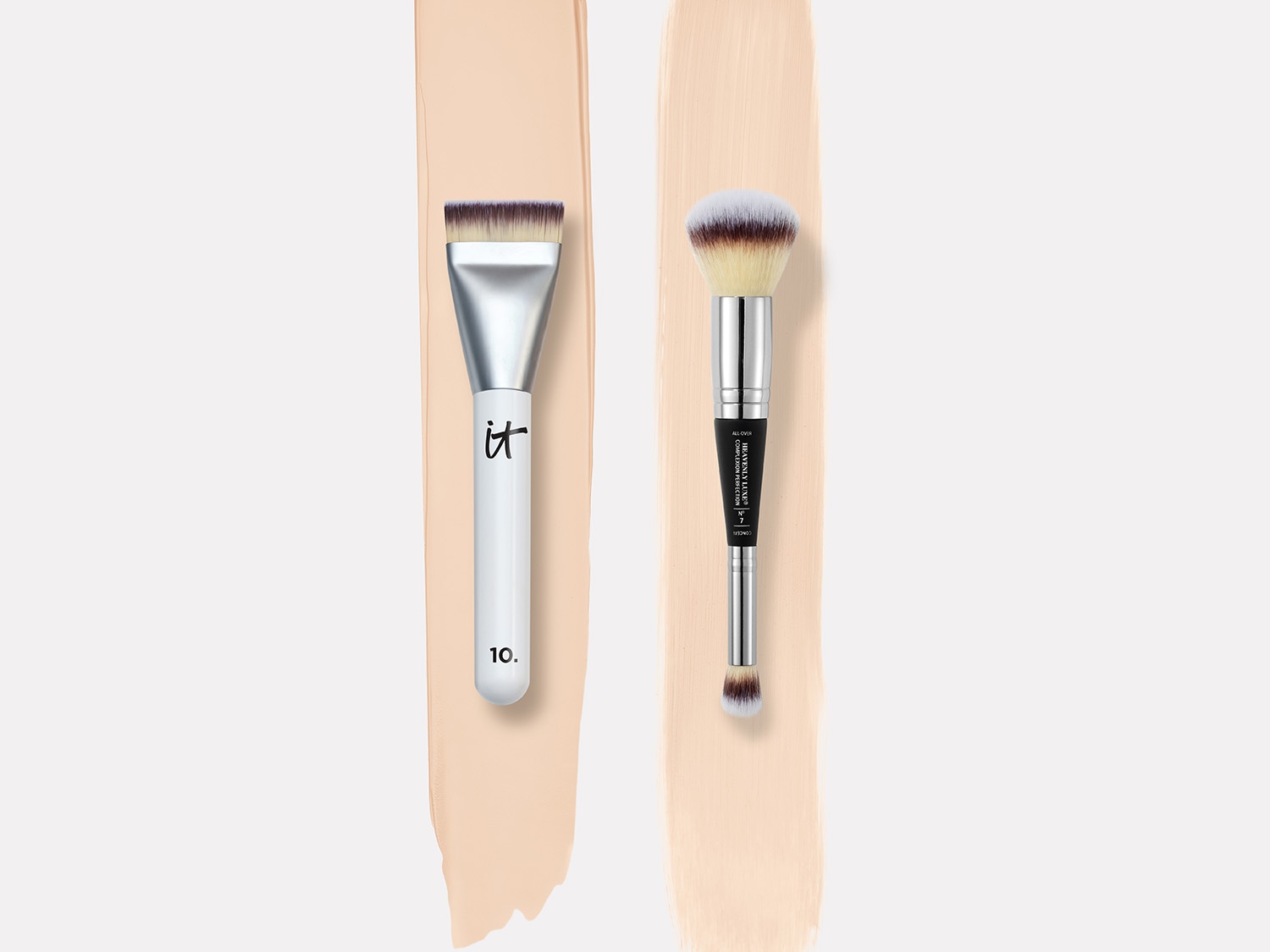 Dwa pędzle do makijażu IT Cosmetics leżą obok siebie na tle beżowych smug, jeden z białym uchwytem i płaską główką, a drugi z srebrnym uchwytem i puszystą główką.