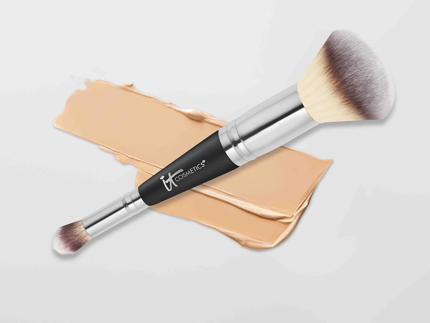 Pędzel do makijażu IT Cosmetics leży na próbce beżowego podkładu, prezentując swoje miękkie włosie i ergonomiczny uchwyt.