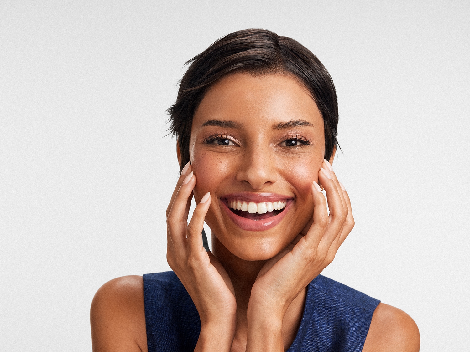 Une femme souriante, les mains sur les joues, rayonne de bonheur avec ses cheveux courts et son teint éclatant.