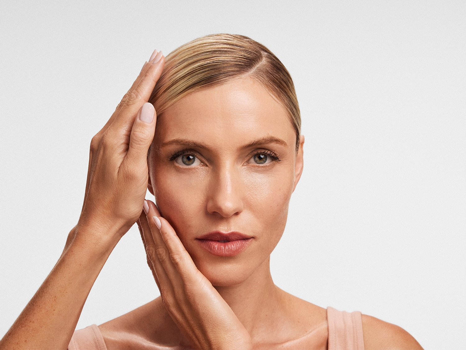 Portrait d'une femme blonde aux yeux clairs, les mains doucement posées sur son visage, peau nette et lèvres rosées.