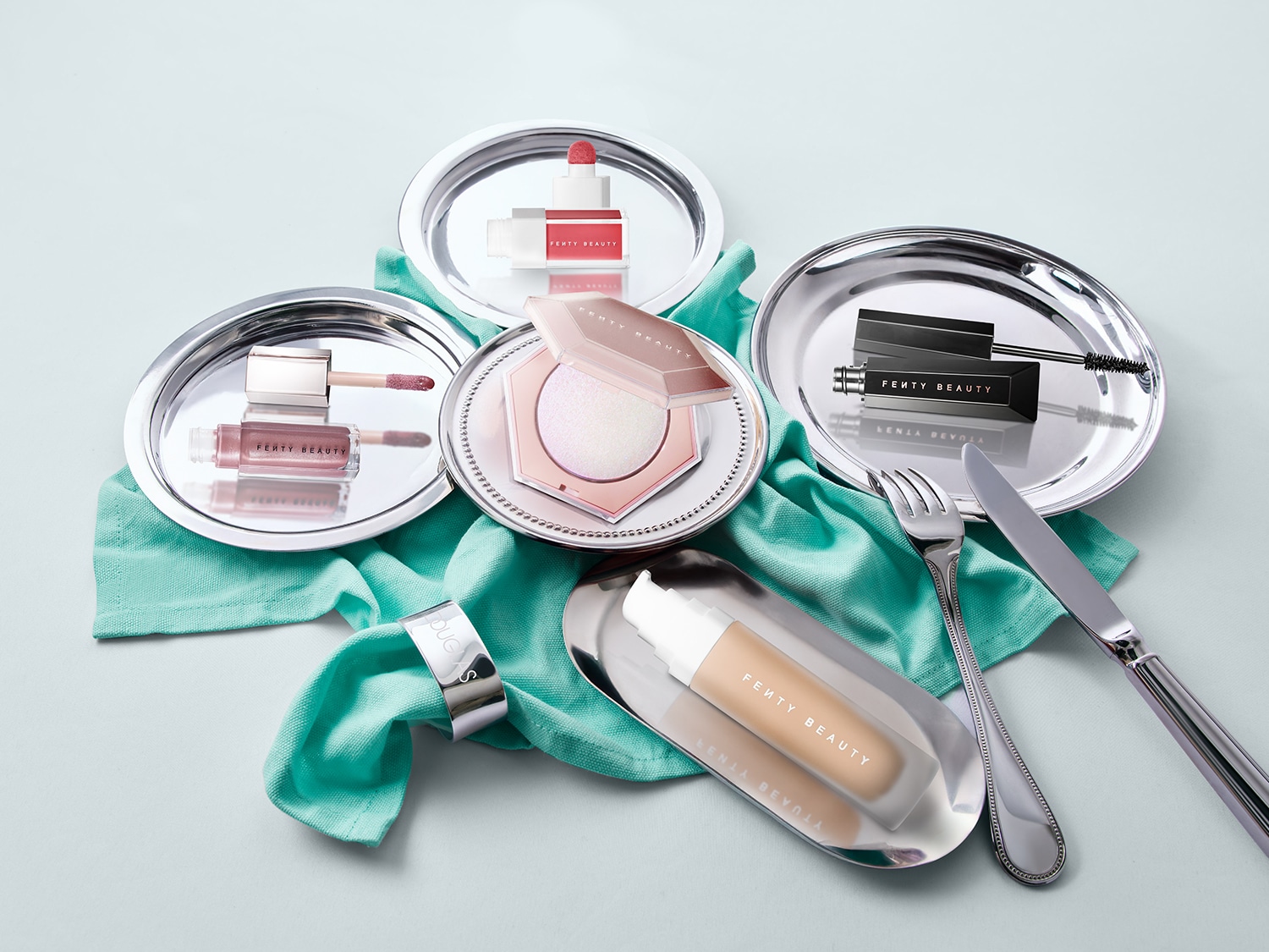 Découvrez une sélection de produits Fenty Beauty présentés sur des plateaux argentés, comprenant des brillants à lèvres roses, un illuminateur nacré, un mascara noir et un fond de teint beige.