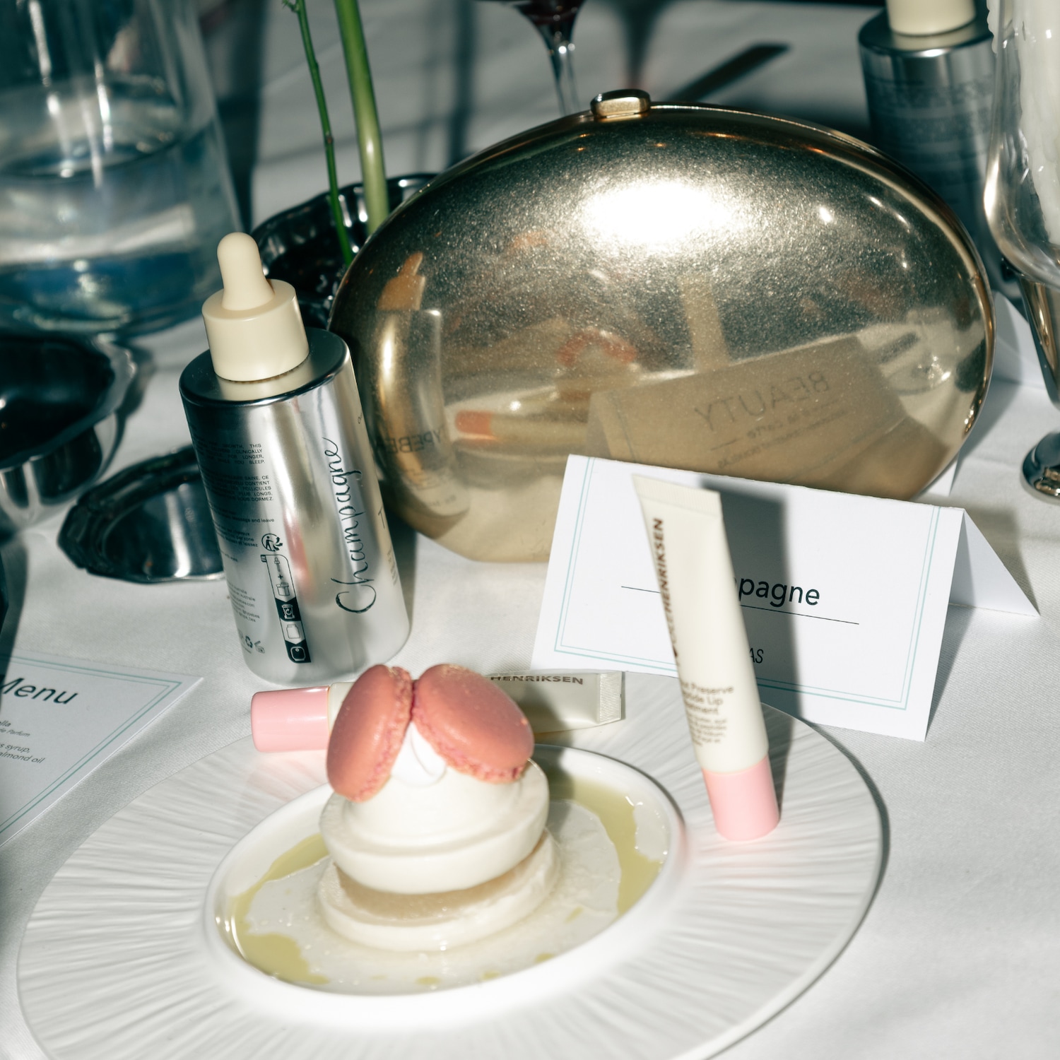 Op een witte tafel staat een gouden clutch, een zilveren flesje serum, een tube lipverzorging en een dessert met roze macarons.