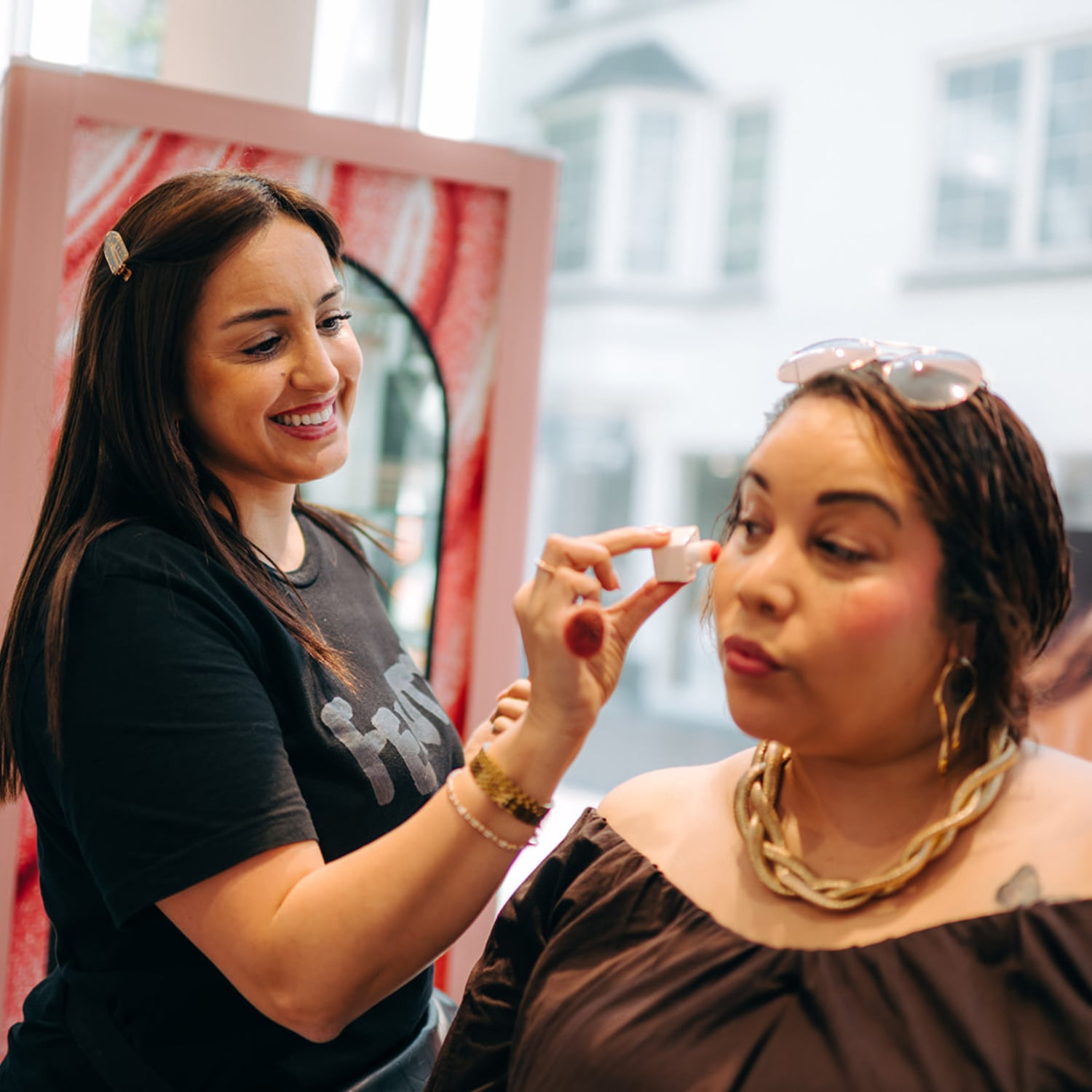 Een visagiste brengt met een klein wit potje make-up aan op de oogleden van een vrouw met een bruine top en gouden ketting.