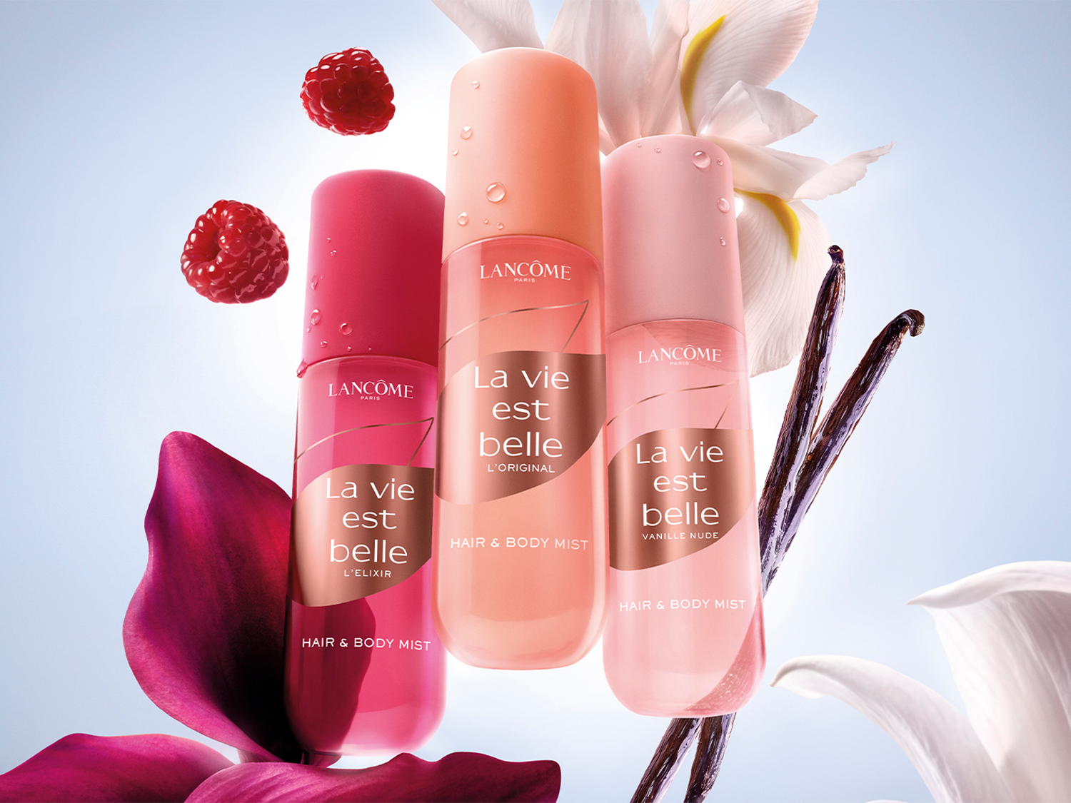 Trzy butelki mgiełki do włosów i ciała Lancôme La Vie Est Belle w odcieniach różu i brzoskwini, otoczone kwiatami i malinami, tworzą elegancką kompozycję.