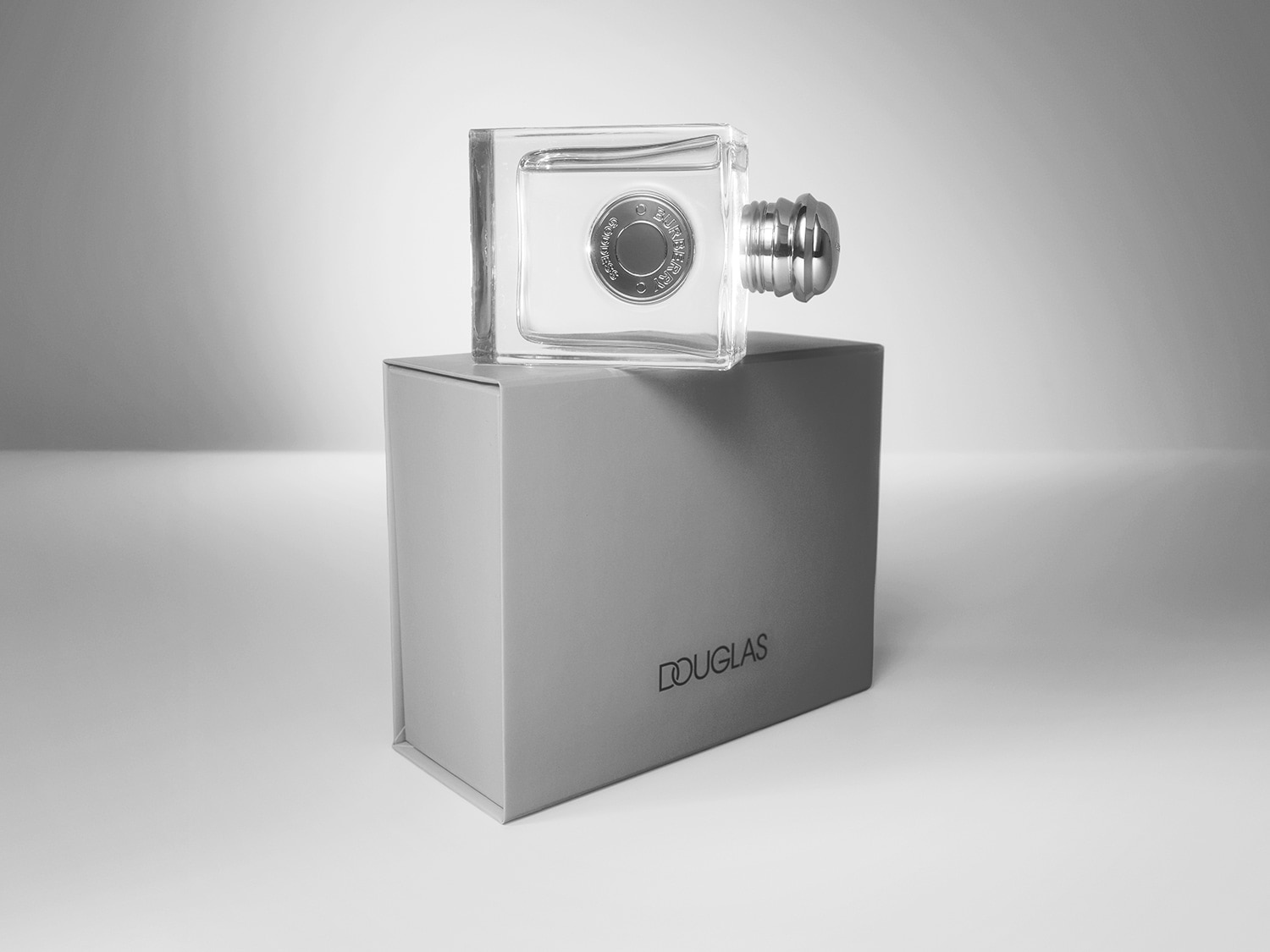 Frasco de perfume transparente con tapón plateado, sobre una caja gris con la marca Douglas, destacando su diseño minimalista y elegante.