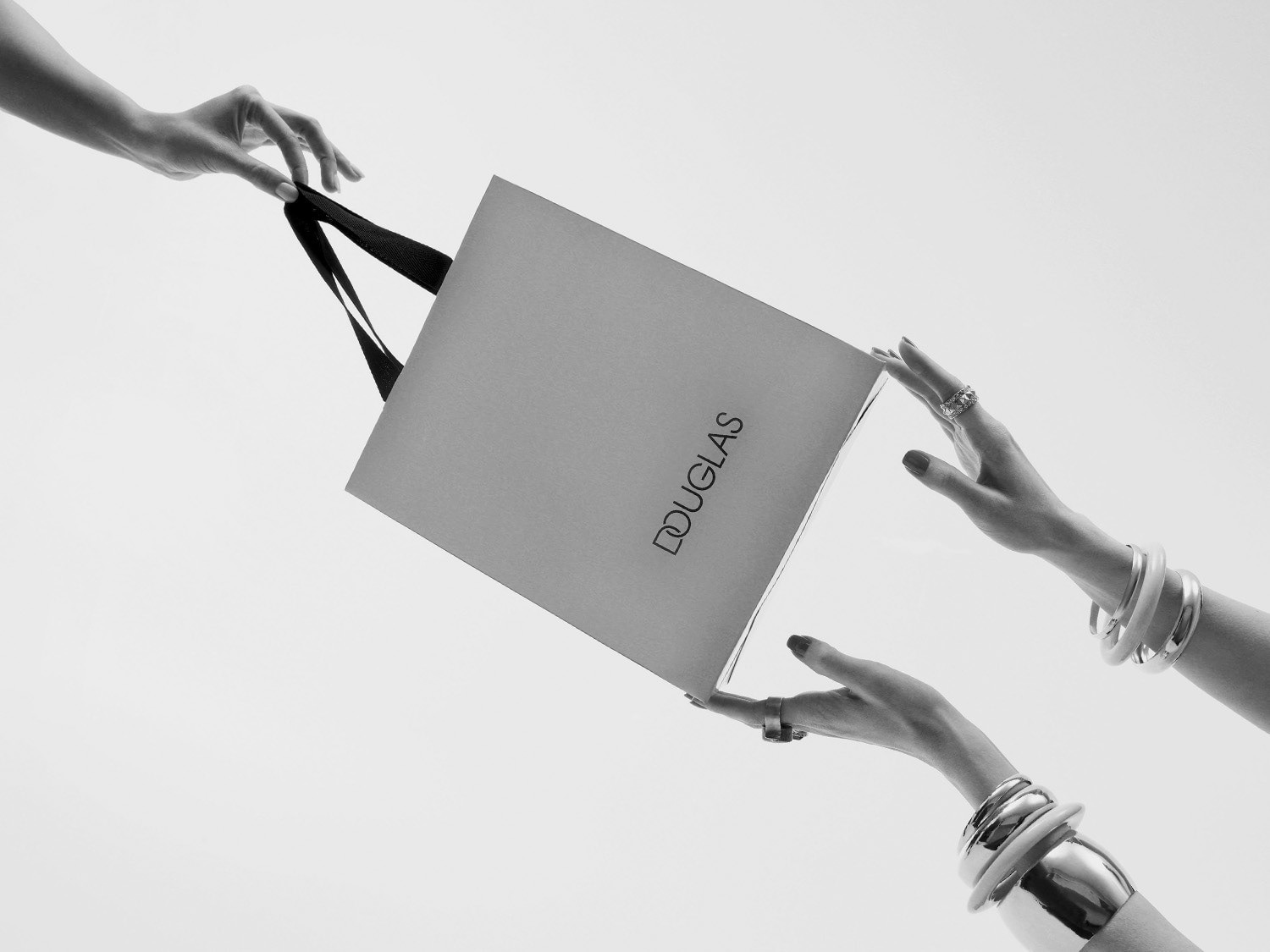En la imagen en blanco y negro, dos manos adornadas con pulseras y anillos sostienen una bolsa de compras de Douglas, transmitiendo una sensación de lujo y elegancia.