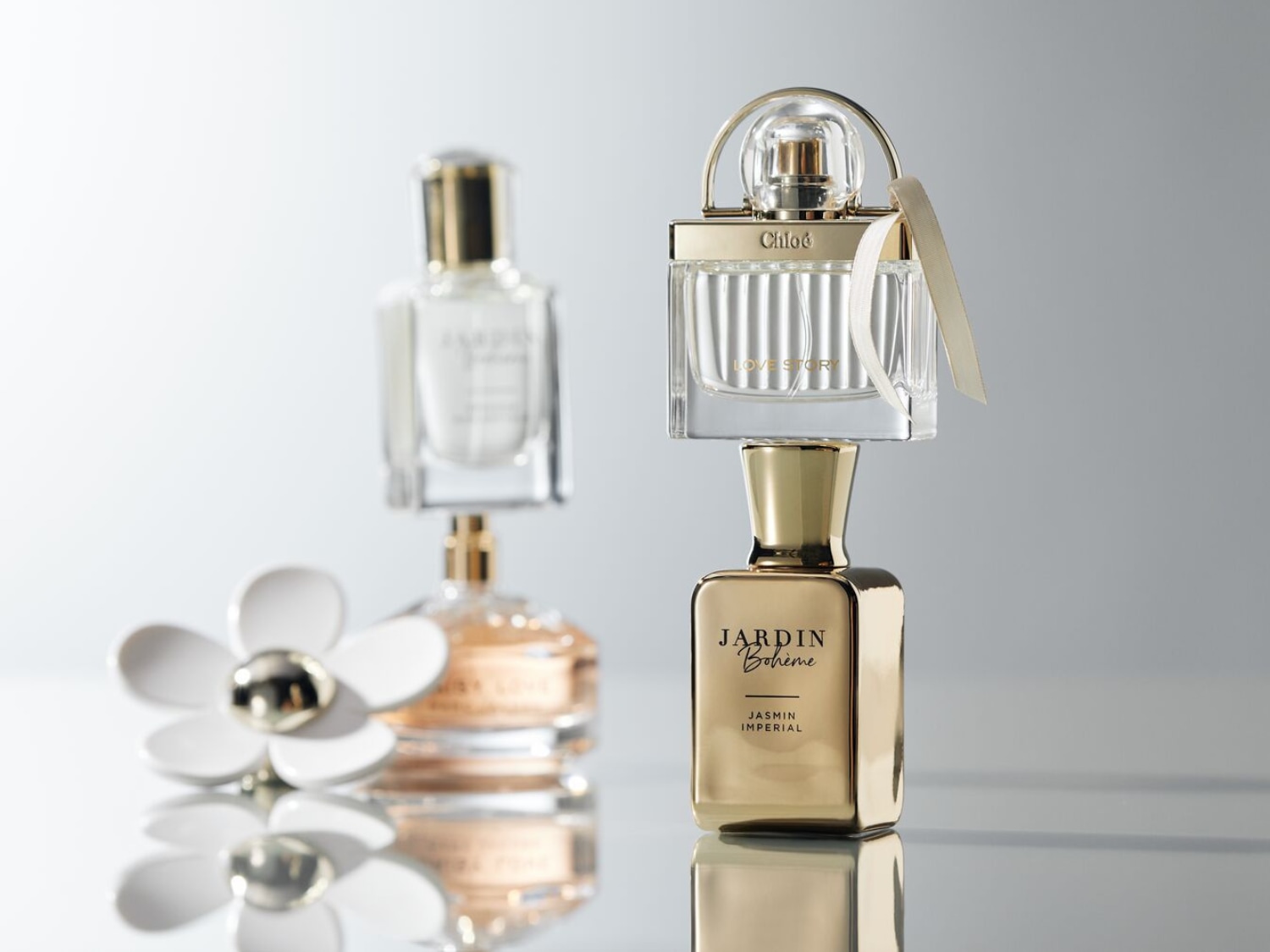 Dwa flakony perfum stoją na błyszczącej powierzchni, z czego jeden to złoty flakon "Jardin Boheme Jasmin Imperial", a na nim przezroczysty flakon "Chloe Love Story" ze złotym uchwytem i beżową wstążką.