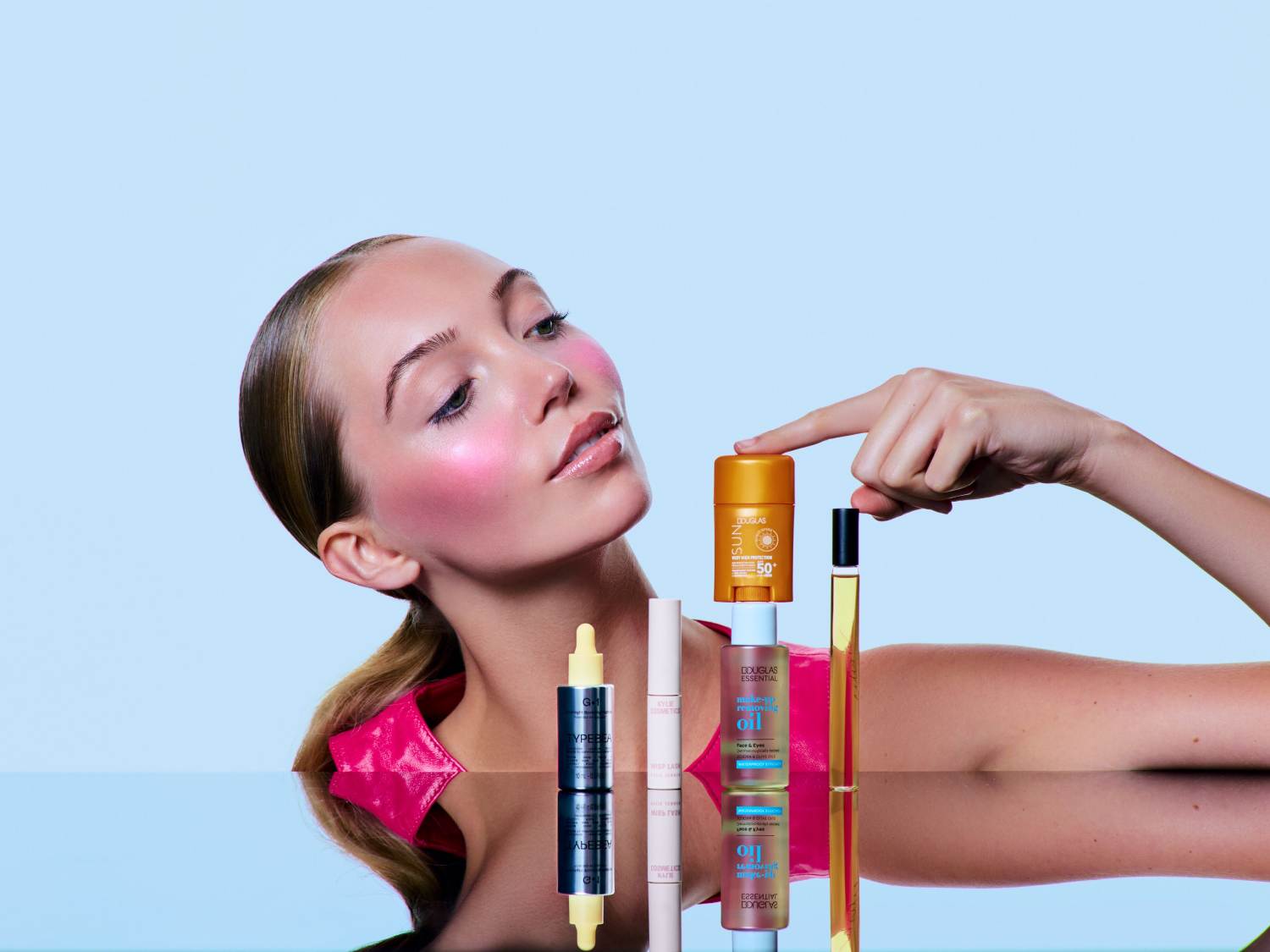 Na jasnoniebieskim tle, kobieta z różem na policzkach prezentuje serię produktów Douglas: olejek, krem z filtrem SPF 50 i serum do twarzy, odbijające się w błyszczącej powierzchni.