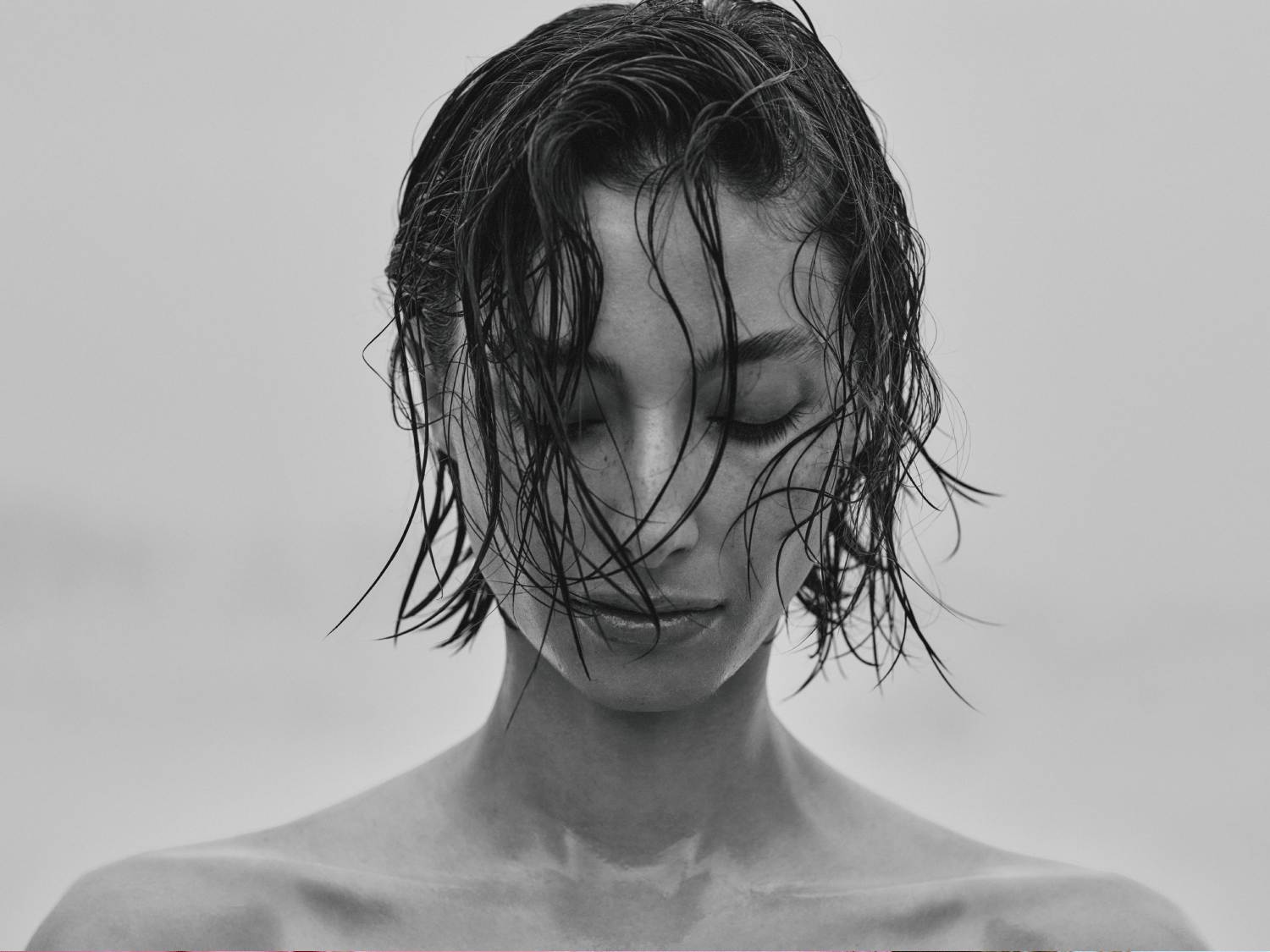 Czarno-białe zdjęcie kobiety z mokrymi włosami opadającymi na twarz i zamkniętymi oczami, emanujące spokojem i naturalnym pięknem.