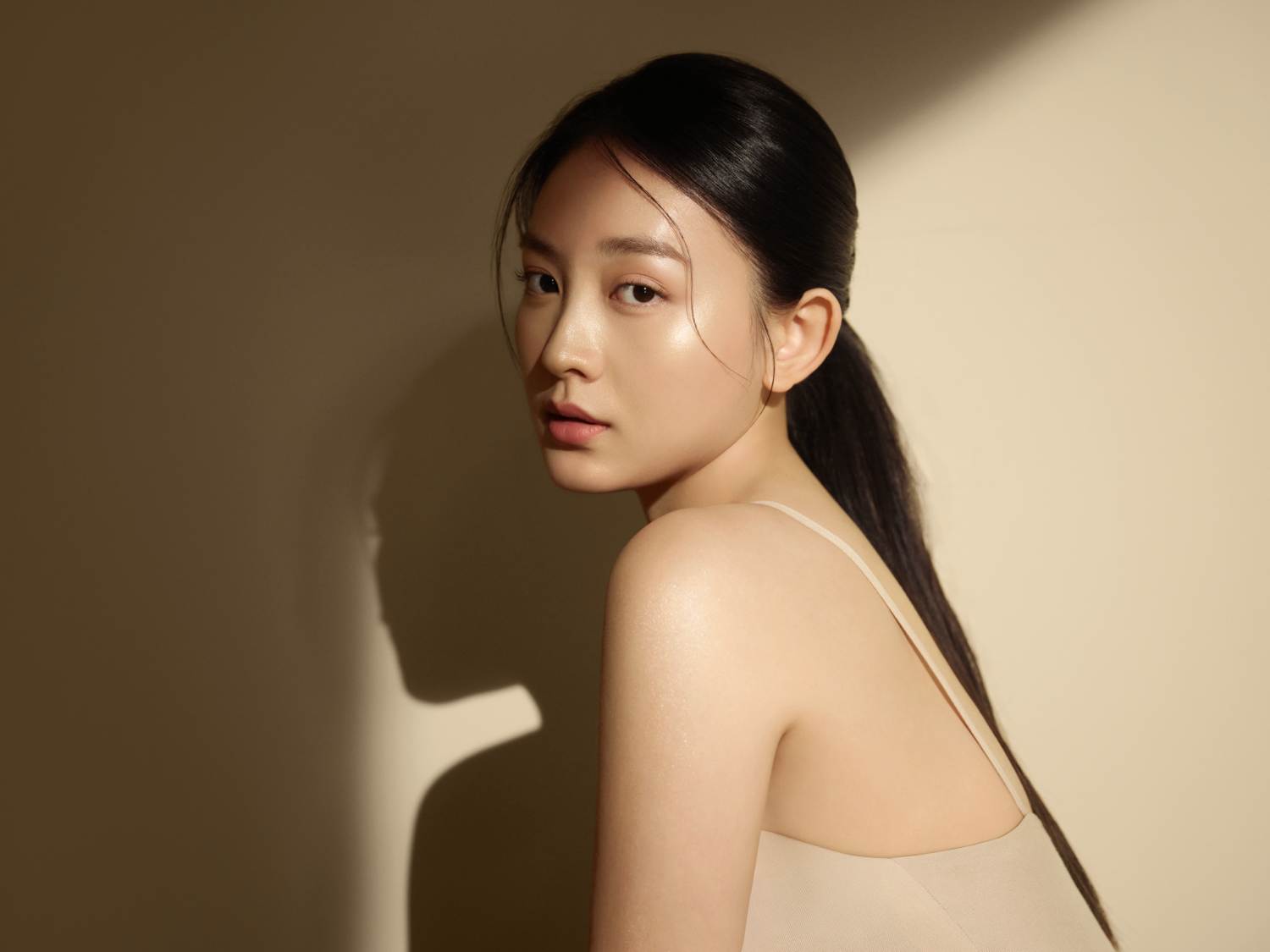 Portret kobiety z czarnymi włosami zaczesanymi do tyłu w kucyk, z delikatnym makijażem i błyszczącą skórą, pozującej na beżowym tle.