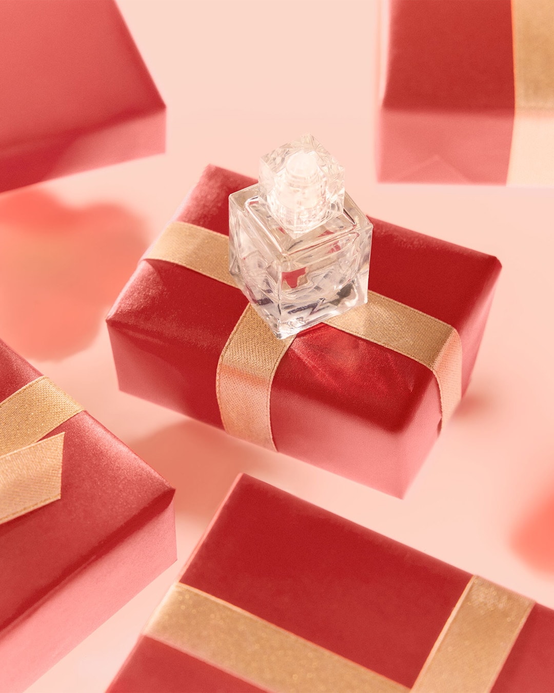 Un parfum transparent repose sur un cadeau rouge orné d'un ruban doré, entouré d'autres cadeaux similaires sur un fond rose doux.