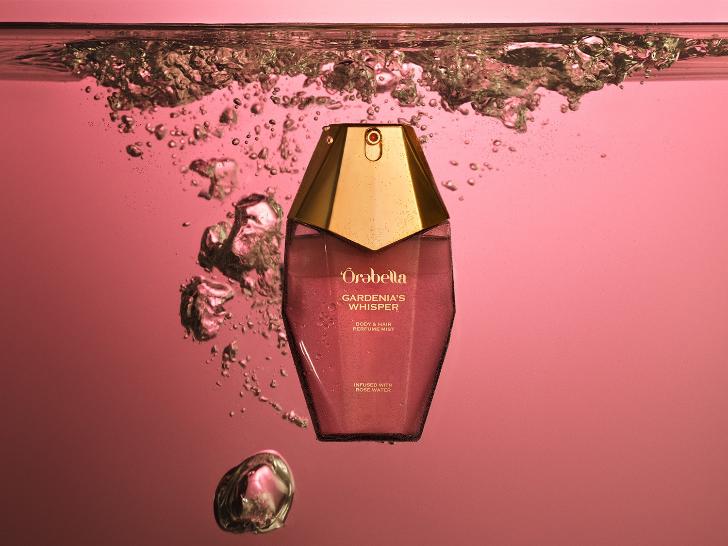 Een roze flesje Oro Bella Gardenia's Whisper body- en haarparfumnevel, met een gouden dop, staat onder water met bubbels eromheen.