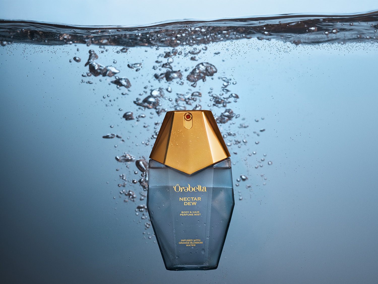 De Orabella Nectar Dew Body & Hair Perfume Mist zit in een blauwe fles met een gouden dop, ondergedompeld in water met bubbels.