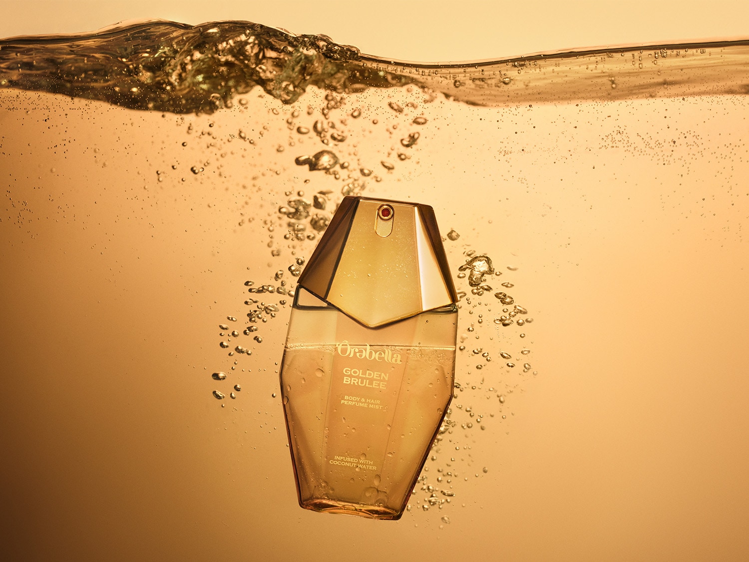 Gouden body- en haarparfumnevel van Orabella, doordrenkt met kokoswater, gehuld in een goudkleurige flacon, zwevend in een gouden vloeistof vol luchtbellen.