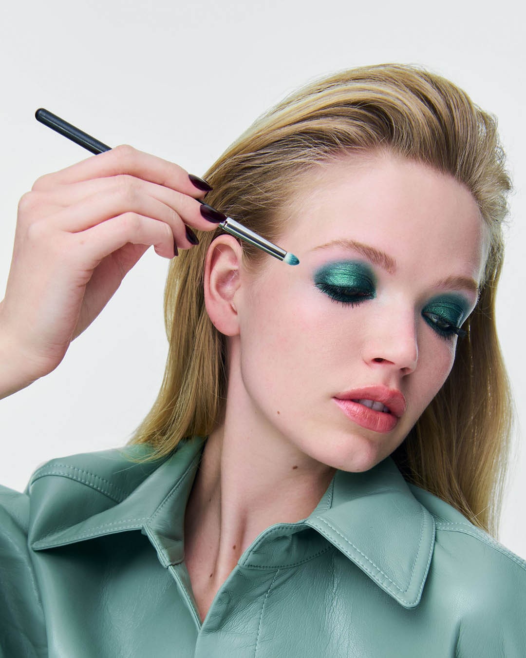 Een vrouw met blond haar en een mintgroen leren overhemd brengt met een kwastje glinsterende groene oogschaduw aan op haar oogleden.