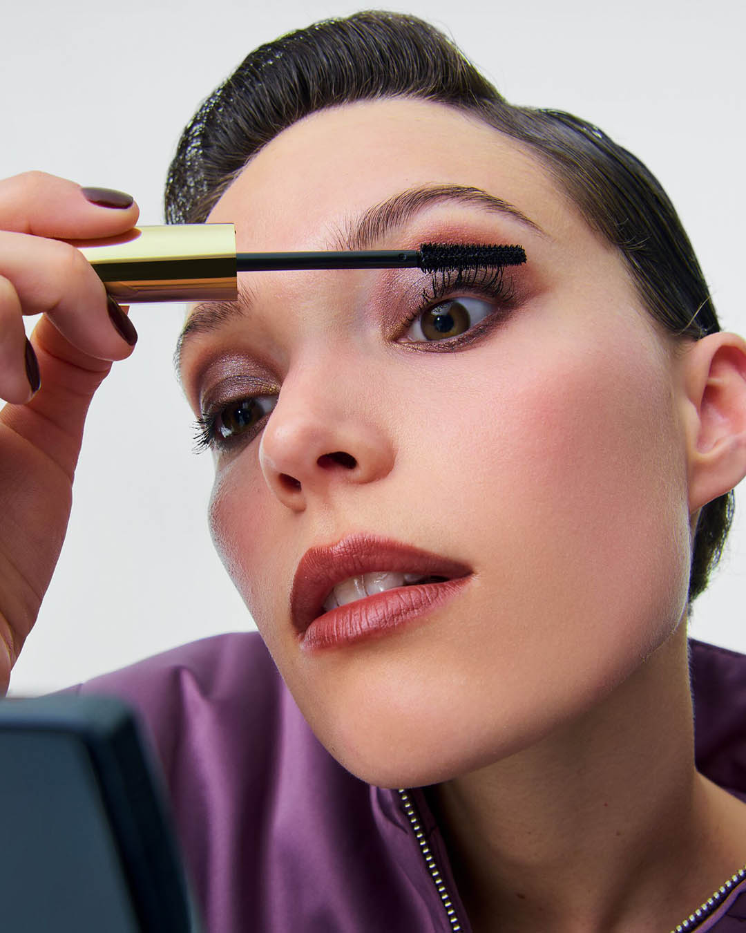 Een vrouw met een paarse jas brengt mascara aan op haar wimpers met een gouden applicator, haar ogen zijn opgemaakt met bruine oogschaduw en haar lippen zijn donkerrood.