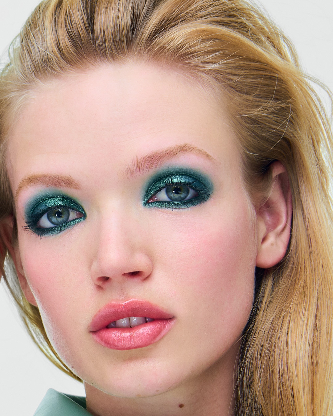 Een close-up van een vrouw met blond haar en een glanzende, smaragdgroene oogschaduw die haar blauwe ogen accentueert, gecombineerd met glanzende roze lippen.