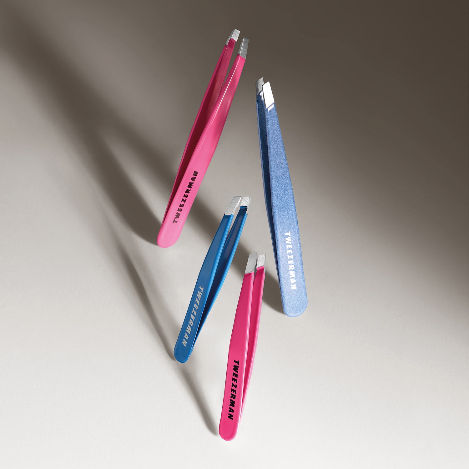 Nahaufnahme von vier Pinzetten der Marke Tweezerman in den Farben Pink und Blau, die auf einer hellen Oberfläche angeordnet sind.
