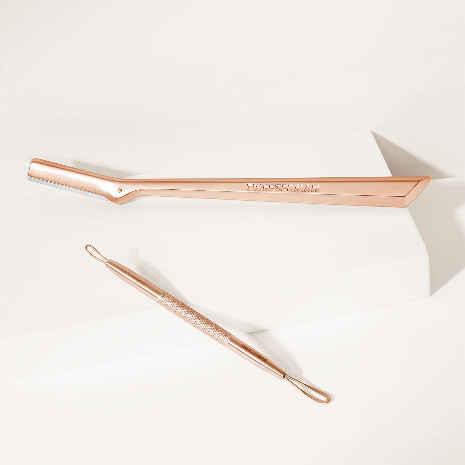 Nahaufnahme von Tweezerman-Tools in Roségold: ein Dermaplaning-Werkzeug mit einer kleinen Klinge und ein Pickel-Extraktor mit einer Schlaufe an jedem Ende.