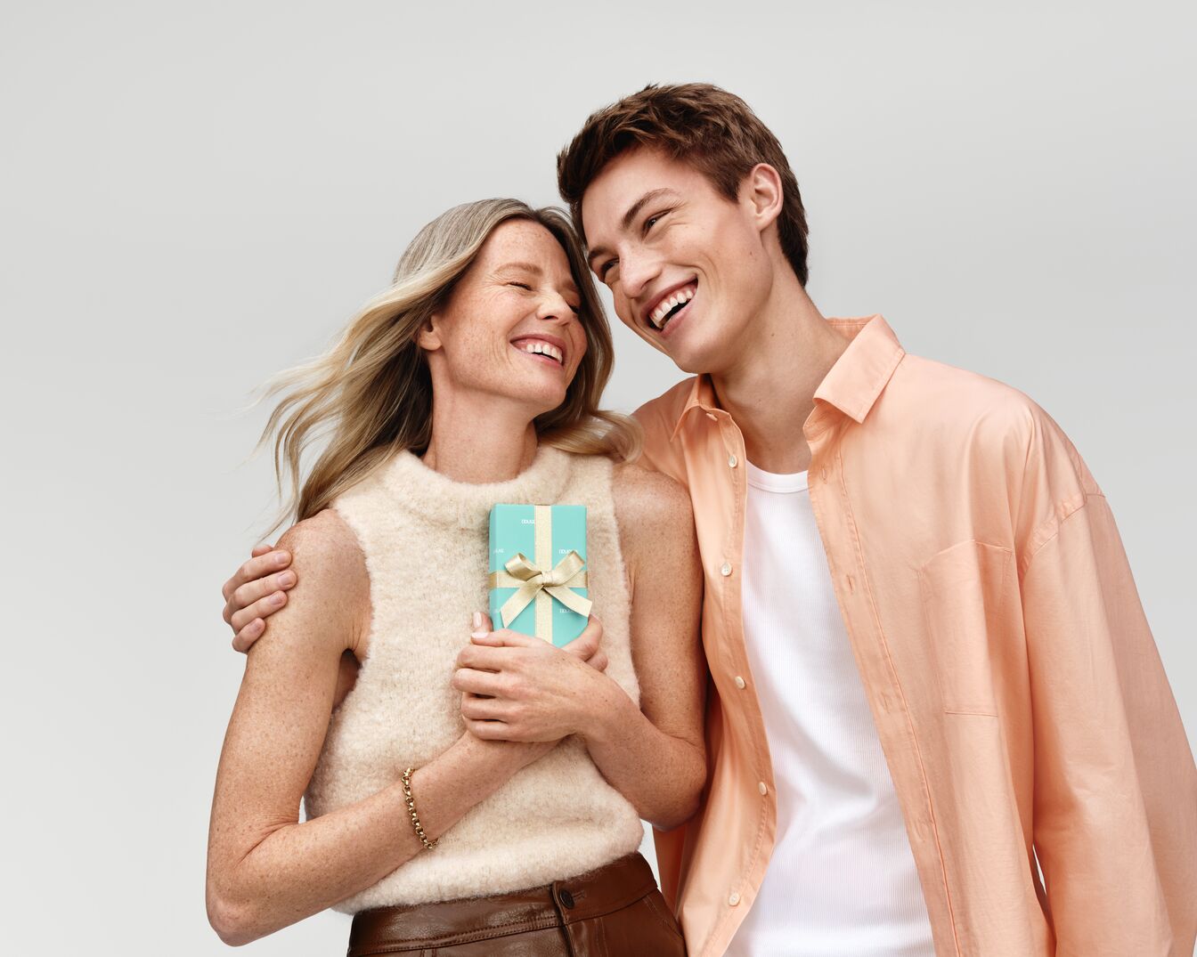 Una donna bionda sorridente riceve un regalo azzurro con un nastro dorato da un giovane uomo, entrambi pieni di gioia.