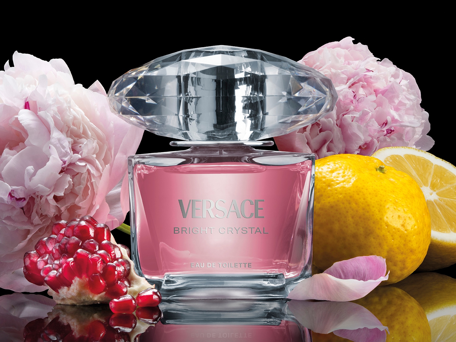 Butelka Versace Bright Crystal z różową zawartością i kryształową nakrętką, otoczona kwiatami piwonii, granatem i cytryną na czarnym tle.