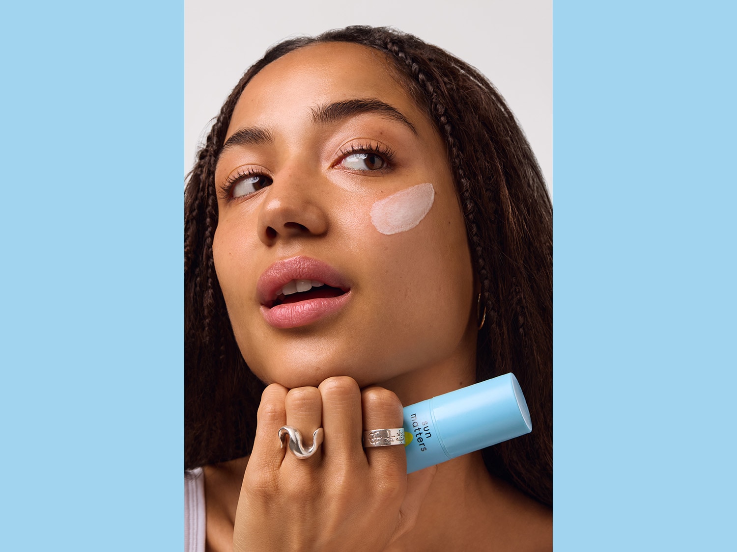 Eine Frau mit Zöpfen hält eine hellblaue Tube Sun Matters und hat eine weiße Creme auf ihrer Wange.