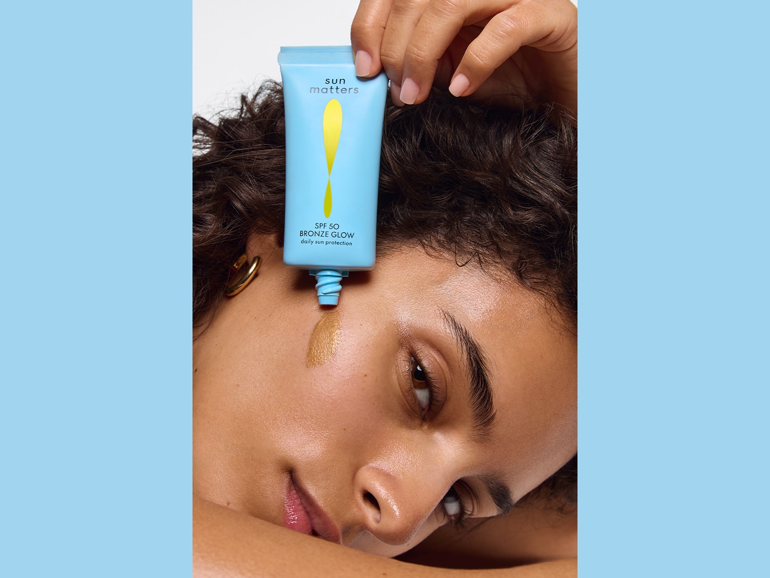 Nahaufnahme einer Frau, auf deren Wange eine bronzefarbene Flüssigkeit aus einer hellblauen Tube mit der Aufschrift Sun Matters SPF 50 Bronze Glow tropft.