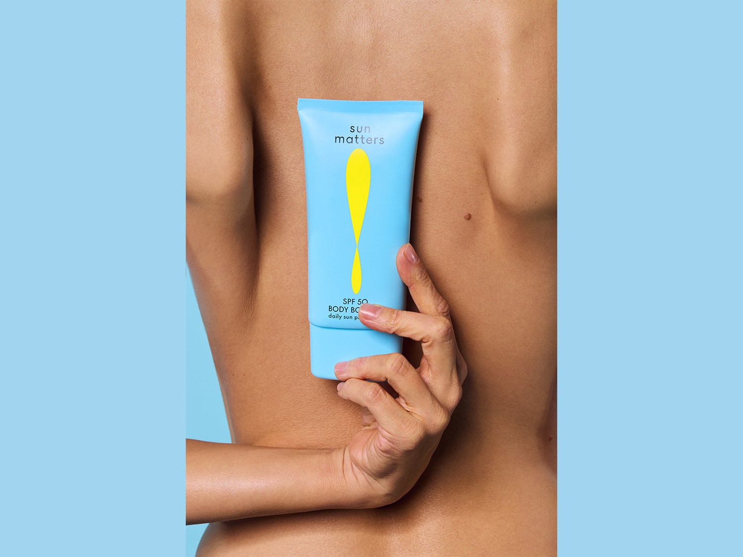 Eine Hand hält eine hellblaue Tube Sun Matters SPF 50 Body Booster Sonnenschutzmittel vor dem Rücken einer Person.