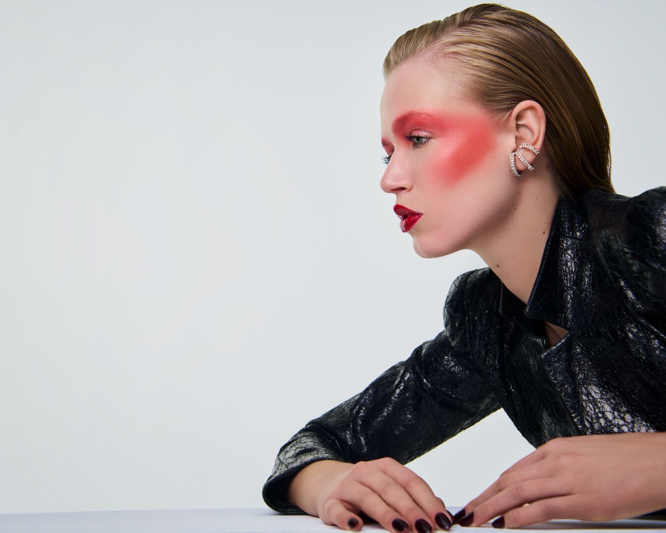 Profil einer Frau mit rotem Rouge, roten Lippen und glänzendem, zurückgekämmtem Haar, die eine schwarze Lederjacke und funkelnde Ohrringe trägt.