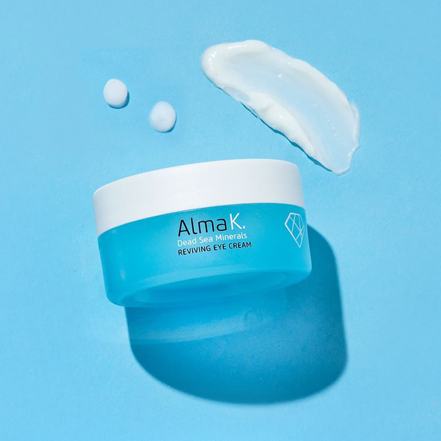 Ecco la crema rivitalizzante per gli occhi Alma K. Dead Sea Minerals, presentata in un barattolo turchese e bianco con un motivo a diamante, accanto a un campione di crema bianca su sfondo azzurro.