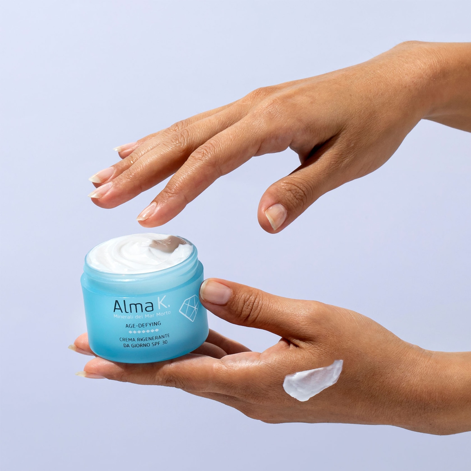 Ecco la crema giorno rigenerante AGE-DEFYING di Alma K, con minerali del Mar Morto e SPF 30, presentata in un vasetto azzurro e applicata sulla mano.