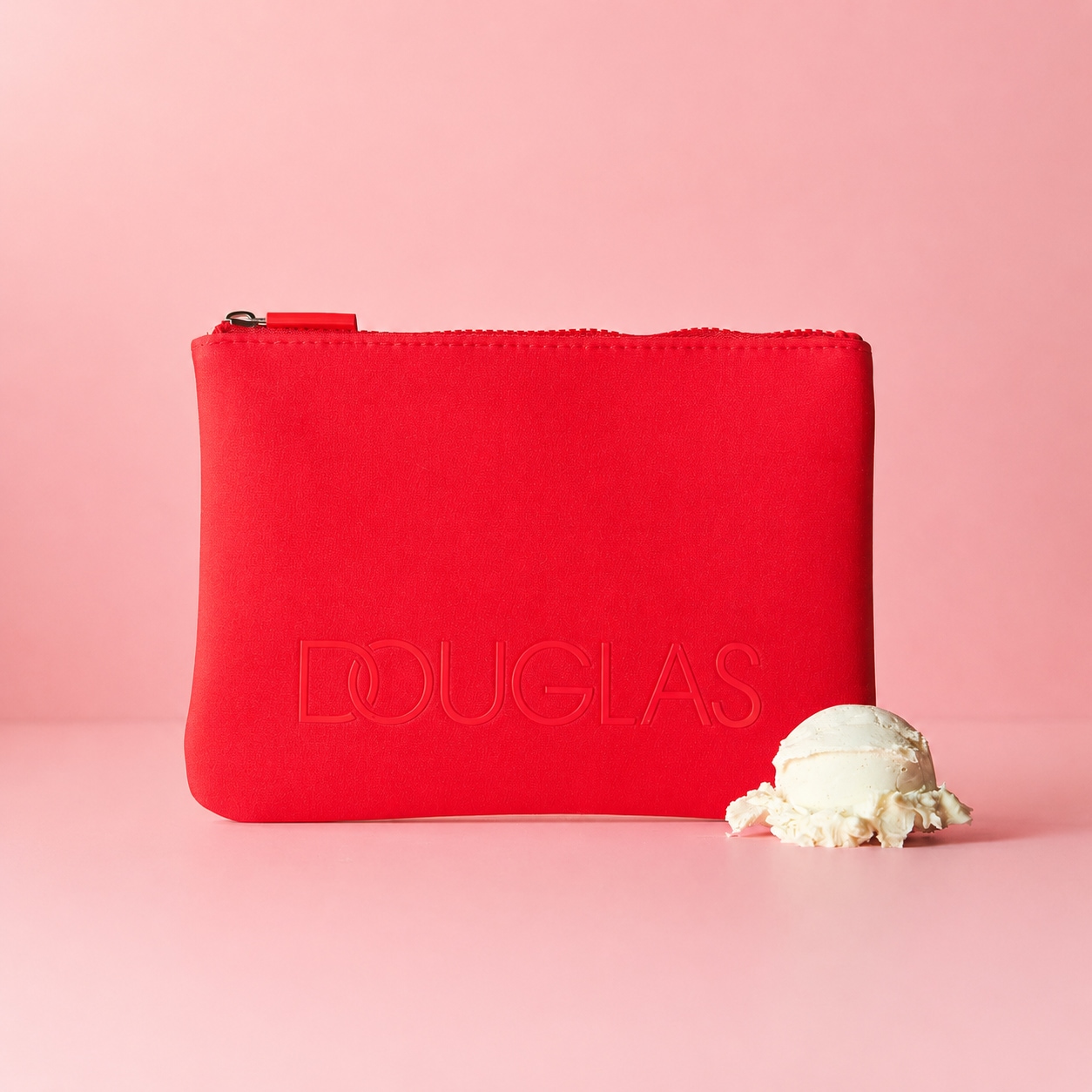 Eine rote Kosmetiktasche von Douglas mit einem Reißverschluss oben und dem Douglas-Logo unten links, daneben eine Kugel Vanilleeis.