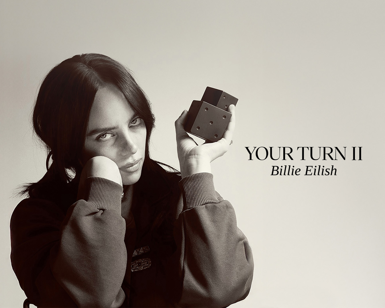 Billie Eilish, cu părul ei închis la culoare, pozează într-un hanorac, ținând în mână două zaruri negre, cu textul "YOUR TURN II Billie Eilish" afișat pe fundal.